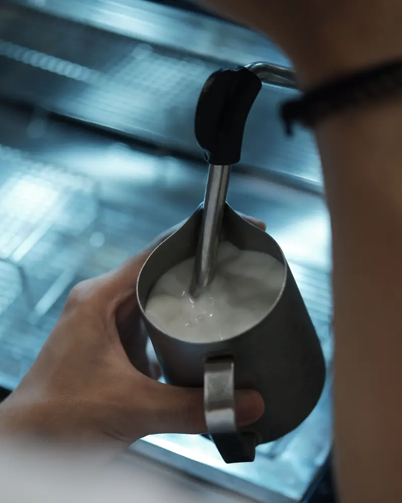 A hand holds a stainless steel milk pitcher under an espresso machine's steam wand while frothing milk.