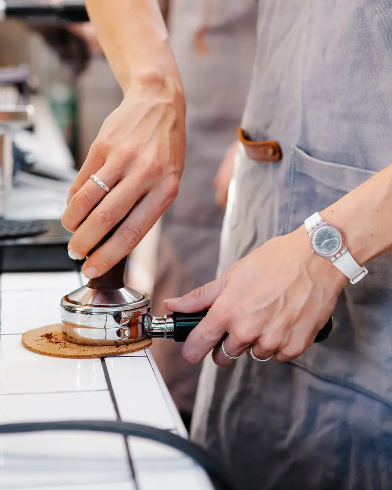 A person in a grey apron tamps ground coffee into a portafilter resting on a cork mat on a white tiled counter.